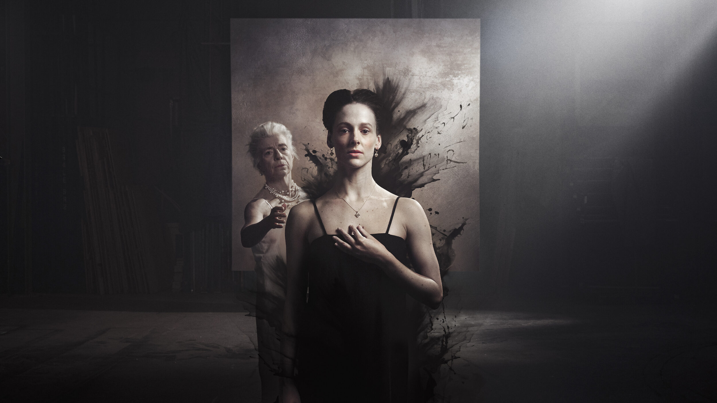 A young dancer faces the camera wearing a black tank top. Her left hand points to her chest. An older woman gestures to her behind her, wearing several pearl necklaces. An abstract black paint design on a white square frames them.