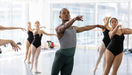 A group of ballet dancers pose in tendu croisé derriere during a ballet class in a large dance studio.