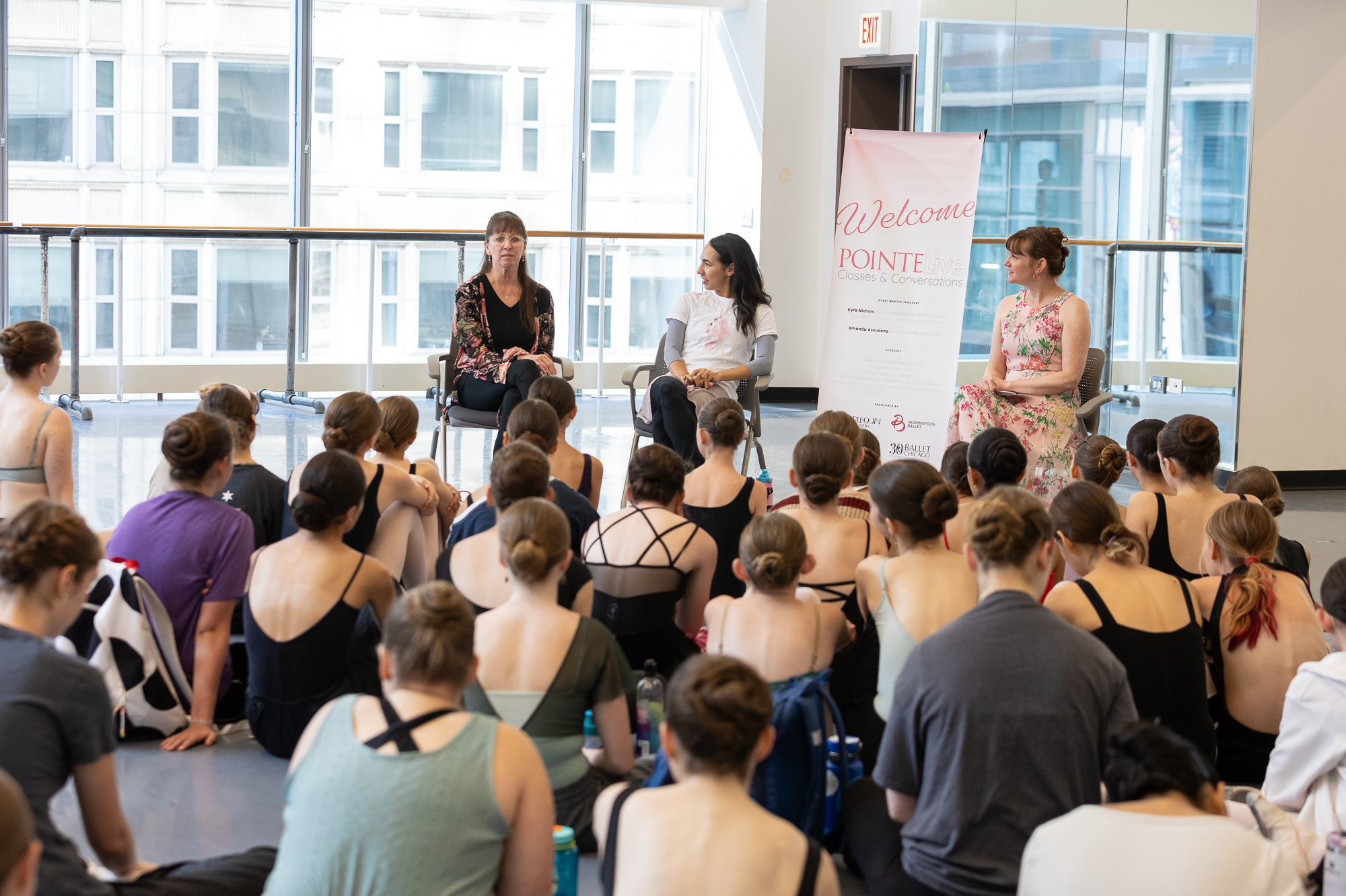 Three women sit in chairs in a large dane studio, facing a large group of tween and teenage ballet students who sit on the floor and listen.