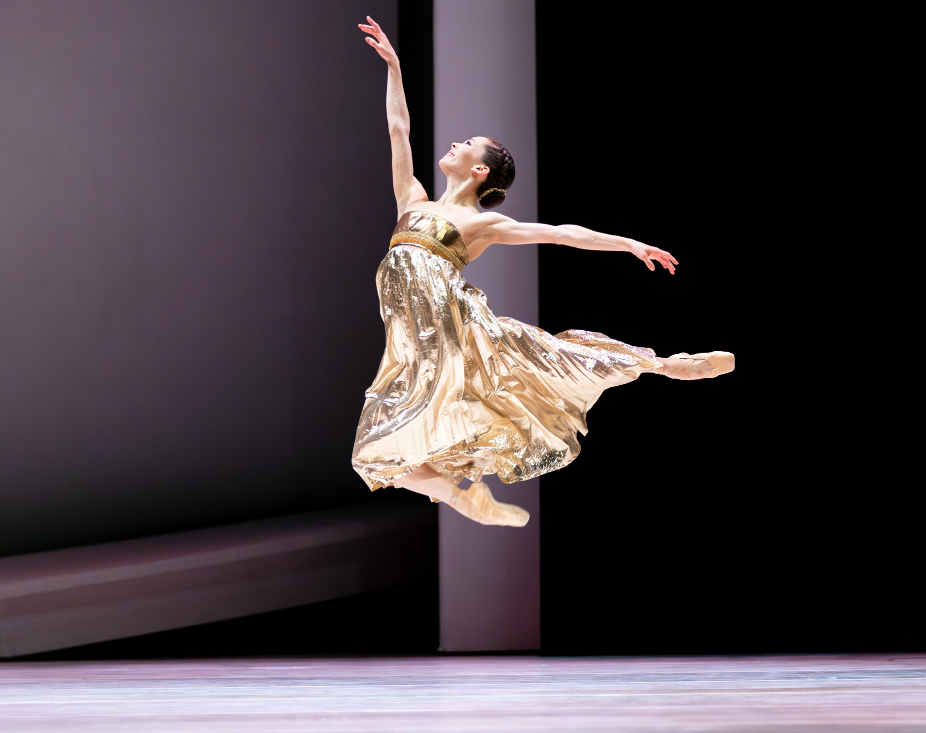 Clara Ruf Maldonado jumps up, tucking her right leg underneath her and extending her left leg back as she faces stage right. She wears a sparkly gold dress and holds her arms in first arabesque, looking up towards her right hand with a large smile.