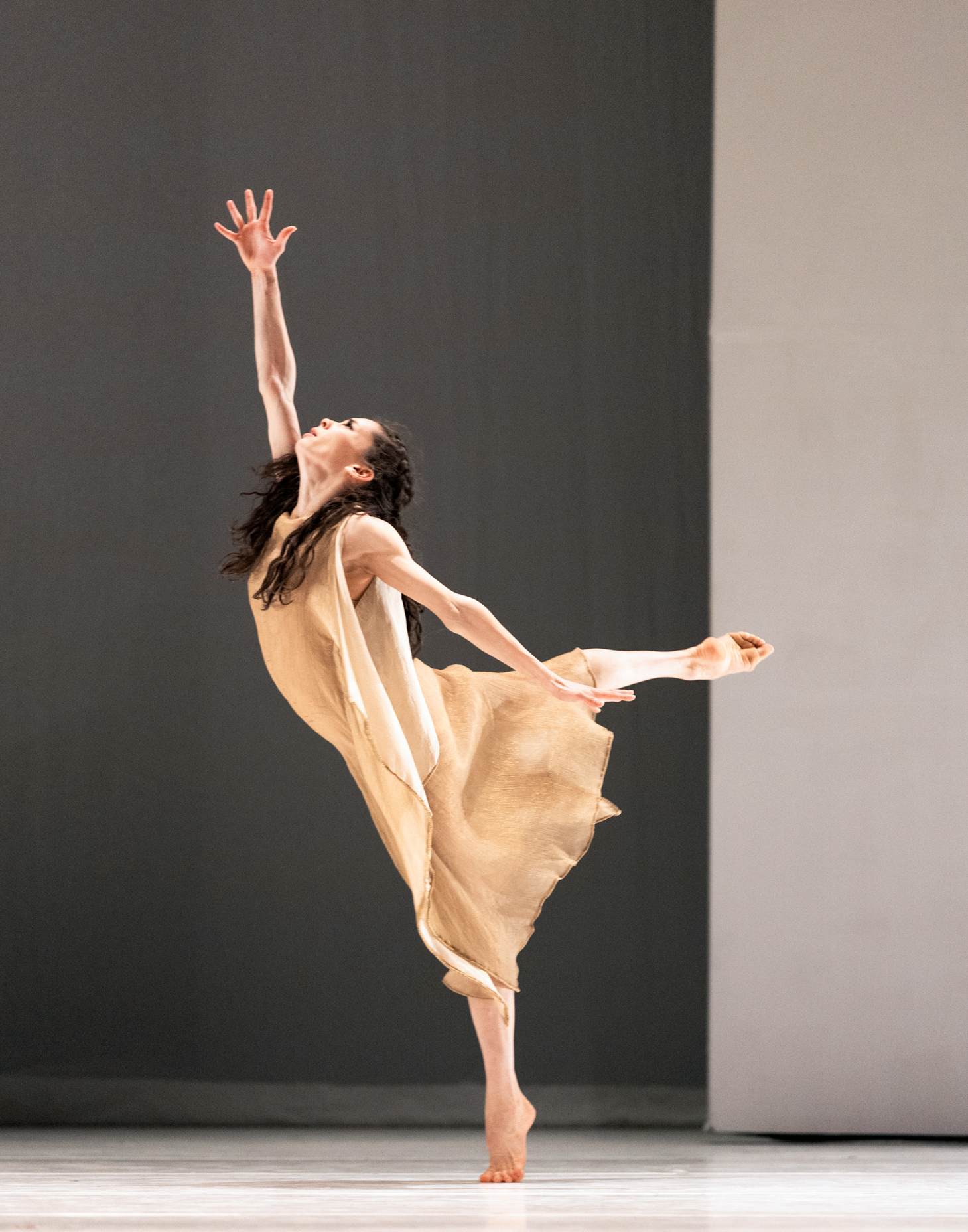 Clara Ruf Maldonado does a croisé arabesque facing stage right. She holds her right arm up, spreading her fingers wide, and looks towards teh ceiling with an anguished look on her face. She is barefoot and wears a filmy tan dress, and her hair down.