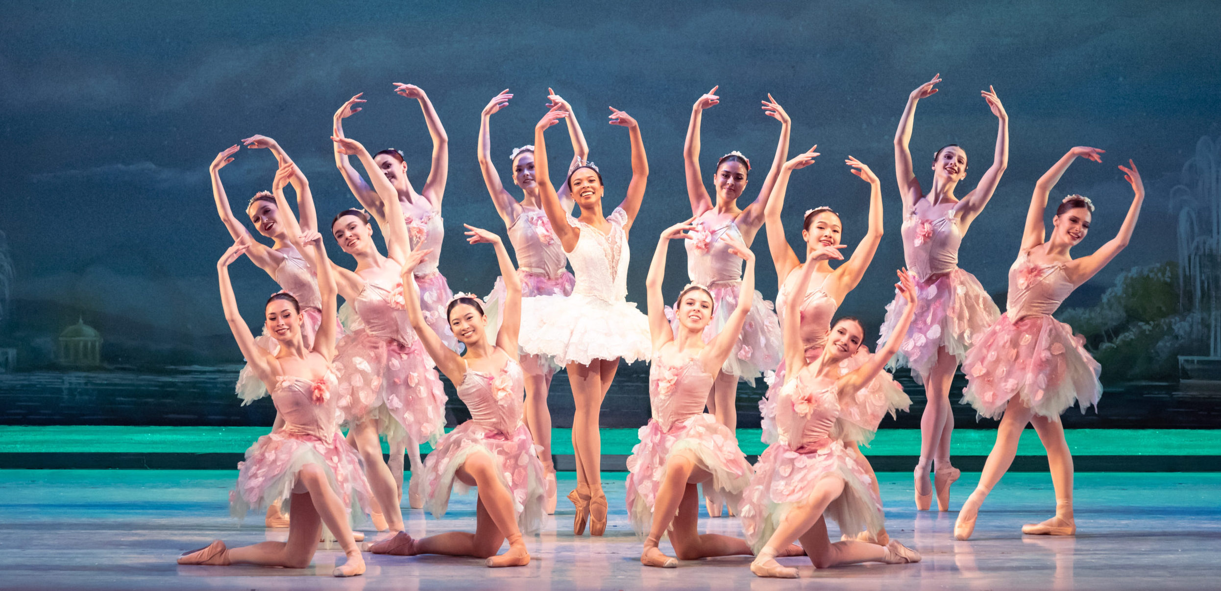 Onstage, Felecia Baker poses in sous-sus on pointe in a white, ruffly tutu with lace details, arms in high fifth. Around her, corps dancers dressed in cherry blossom dresses pose for a final tableau.