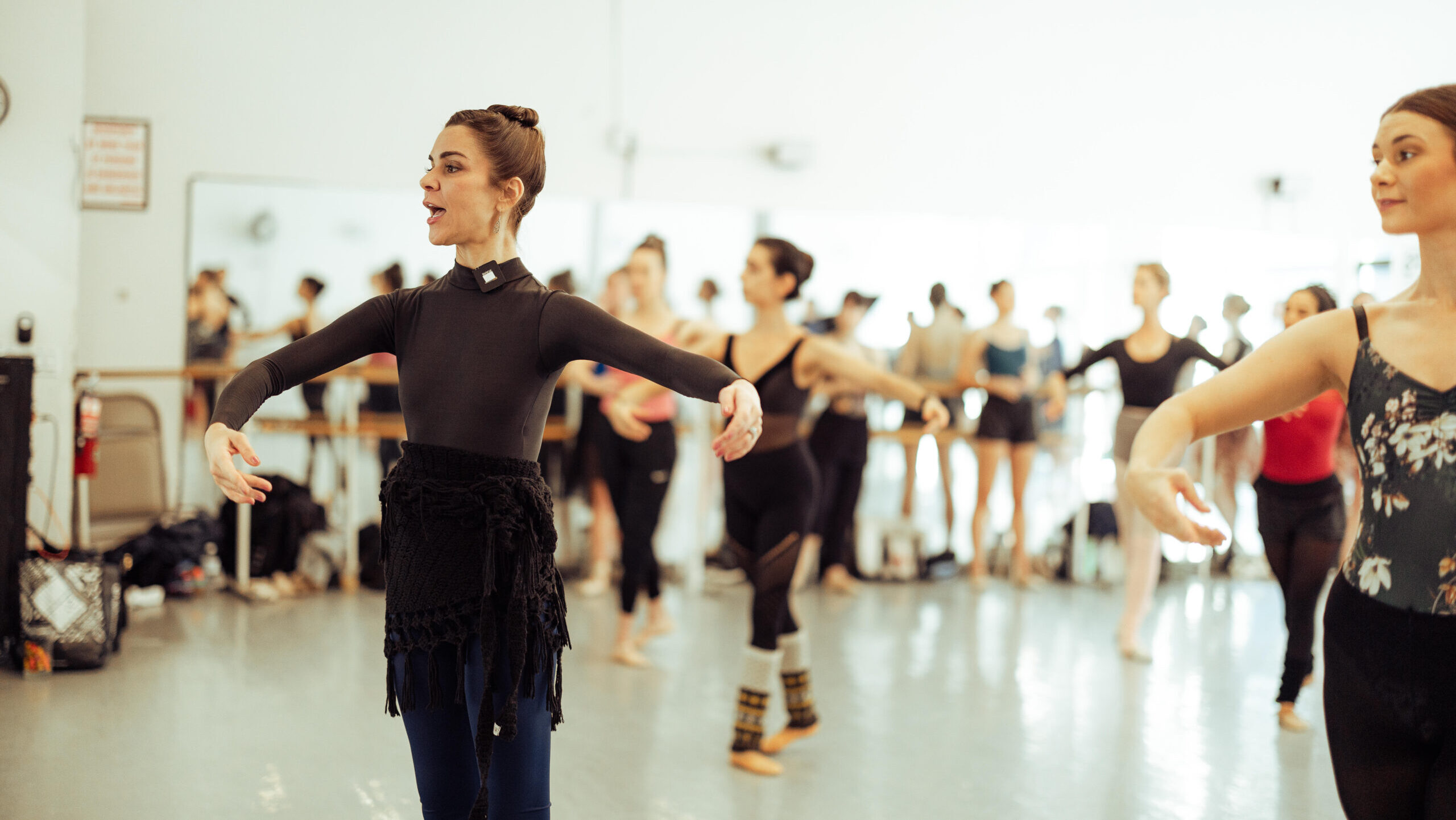 Kate Loh, a tall ballet teacher with brown hair in a high bun and black dancewear, leads a class in a bright dance studio