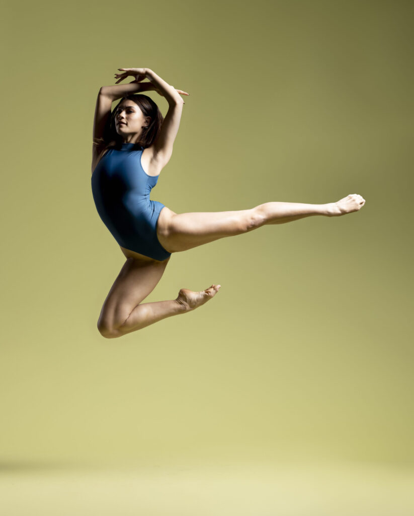 Natalie Taylor jumps. She wears a blue leotard and is against a yellow/green background.