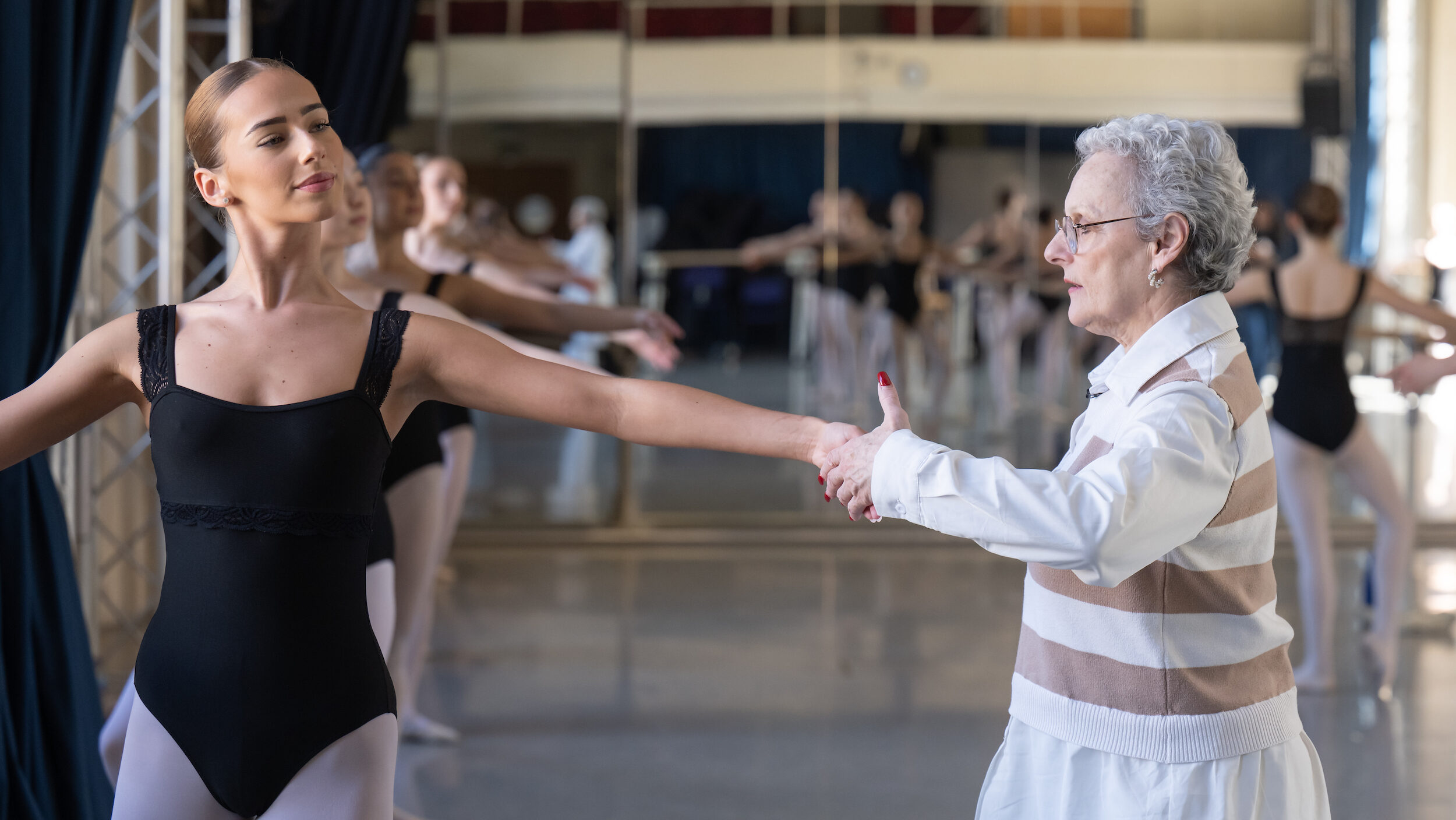 Lynne Charles, wearing a white blouse with a white and tan striped sweater vest over it, adjusts the hand of a ballet student at the barre during a class. All of the dancers wear black leotards and pink tights and shoes.