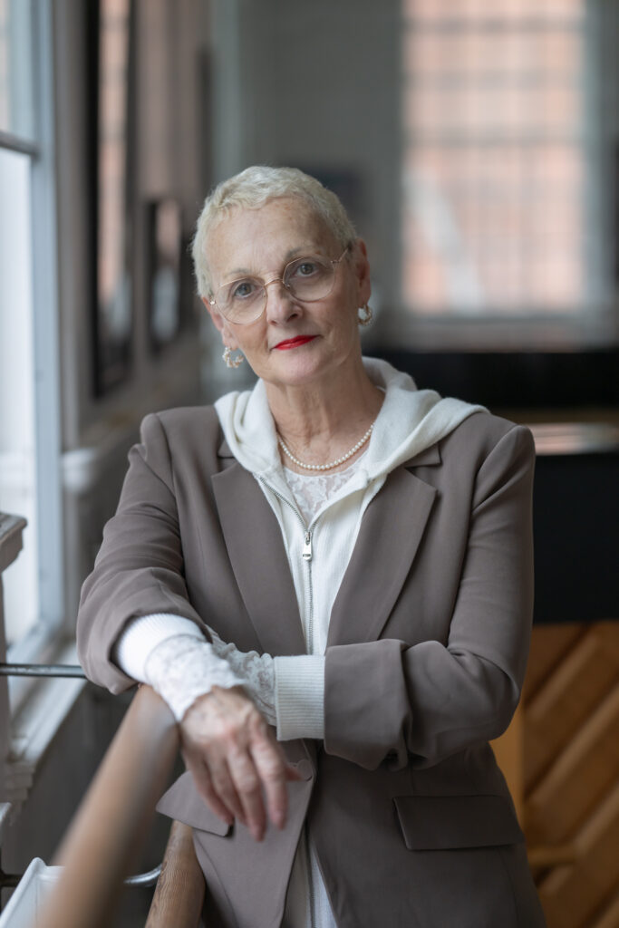 Lynne Charles leans against a ballet barre and looks toward the camera with a slight smile. She wears a tan sport coat over a white top, glasses, and silver earrings.