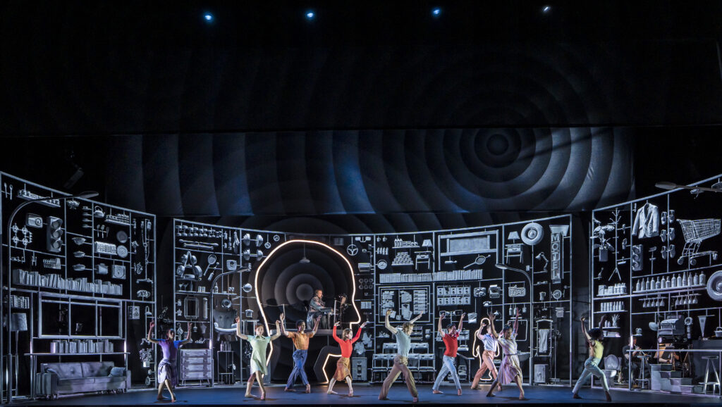 Onstage, a large group of dancers dressed in '60s-style costumes perform in front of a large set piece displaying everyday items and a massive head silhouette in profile.