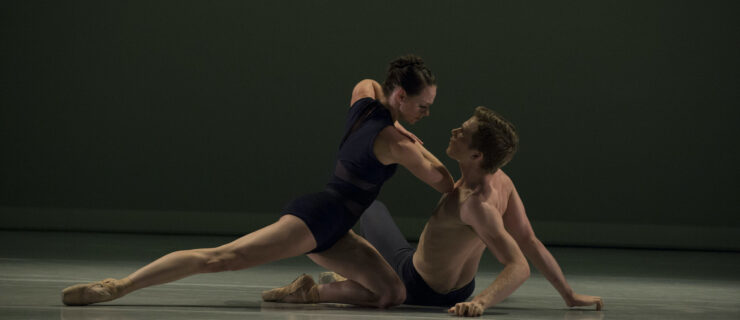 A male and female dancer perform a pas de deux on a darkened stage. The man, in dark tights, sits on the ground as the woman, in a black leotard and pointe shoes, kneels close to him and pushes him down forcefully.