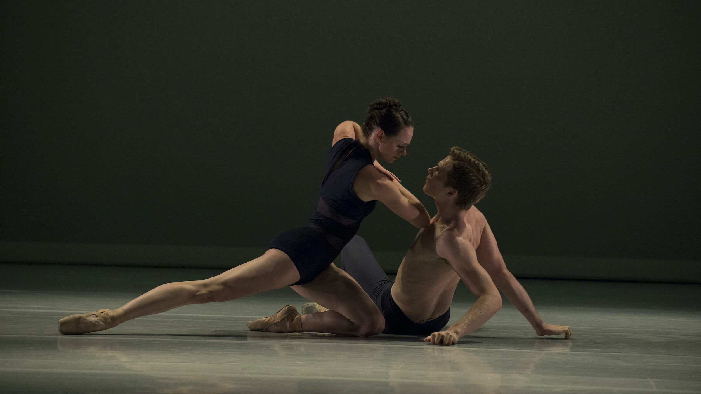 A male and female dancer perform a pas de deux on a darkened stage. The man, in dark tights, sits on the ground as the woman, in a black leotard and pointe shoes, kneels close to him and pushes him down forcefully.