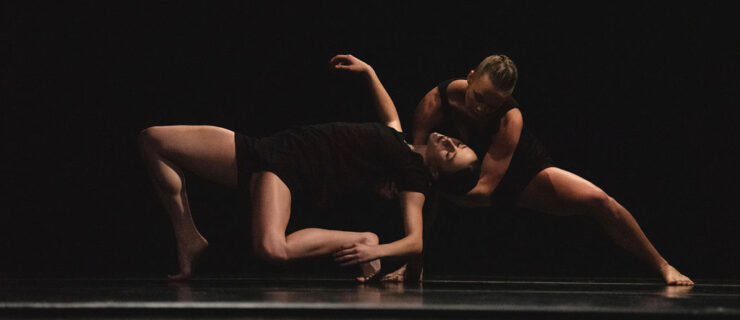 Two barefoot, female dancers perform a contemporary pas de deux on a darkened stage. One dancer bands backwards into a deep hinge, while the other crouches behind her and supports her upper back with her hands.