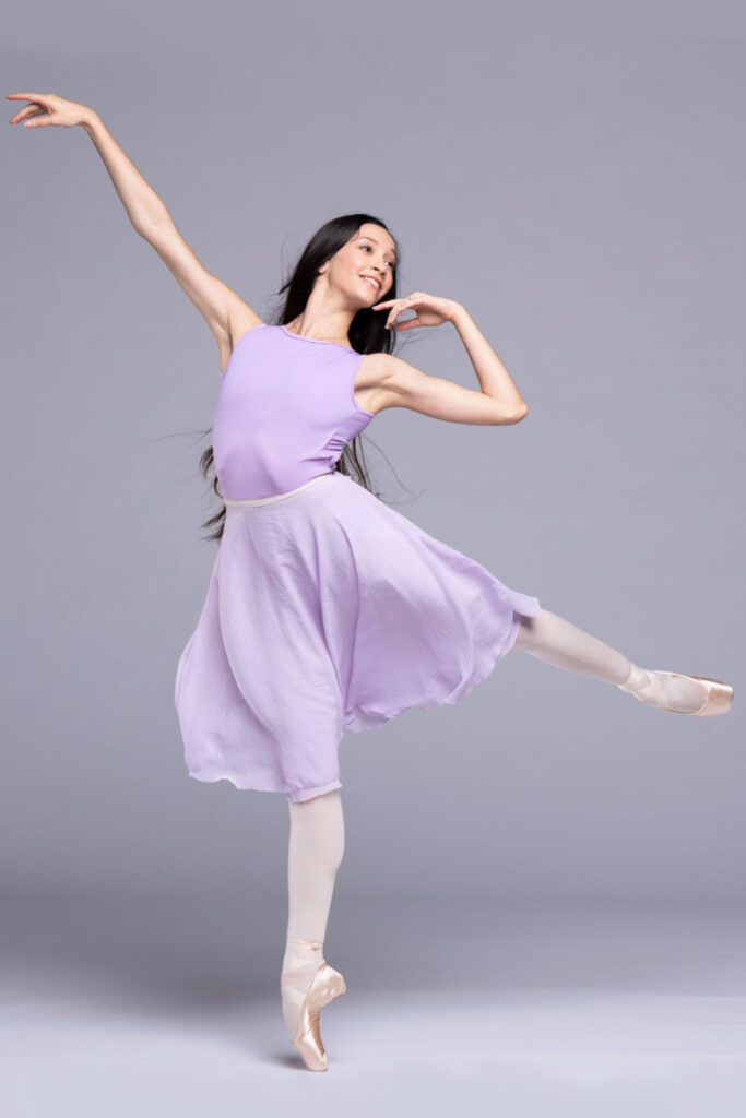 Macarena Giménez, wearing a purple dance dress, pink tights and pointe shoes, poses in a low arabesque on pointe with her left leg up. She looks over her left shoulder, raises her right arm up and pulls her left arm in, brushing her chin with her left hand.