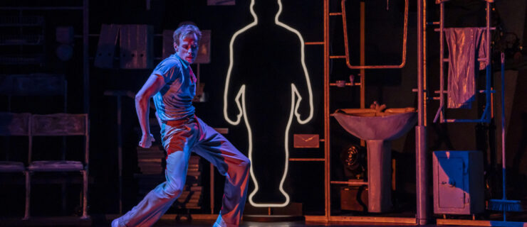 Onstage in vibrant lighting, with a dark background, Ed Watson hinges on forced arch and curves his arm back, looking down and back with an intense expression. He wears slacks and a polo, and behind him is a yellow outline of a man's silhouette.