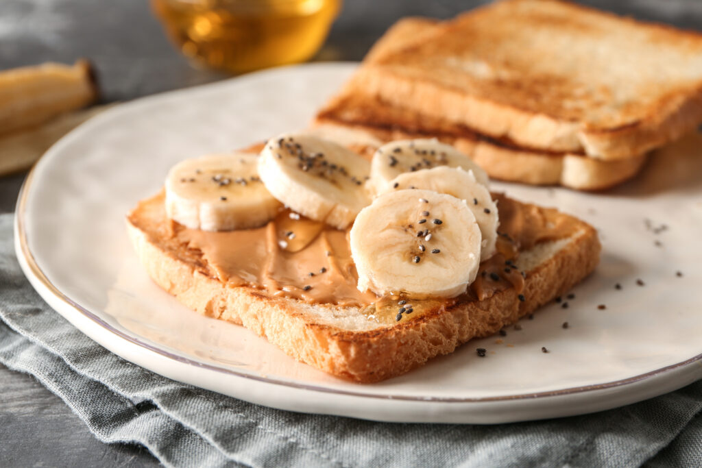 On a table with a cloth napkin, a plate with pieces of toast. The one in front is topped with nut butter, banana slices, and chia seeds.