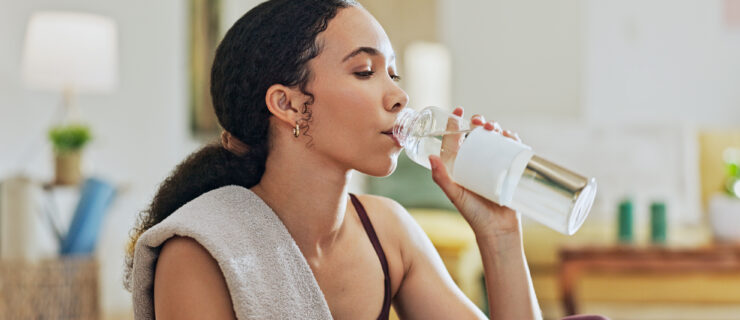 A young woman shown from the shoulders-up, with a sweat towel draped over one shoulder, drinks from a large water bottle after a tough workout.