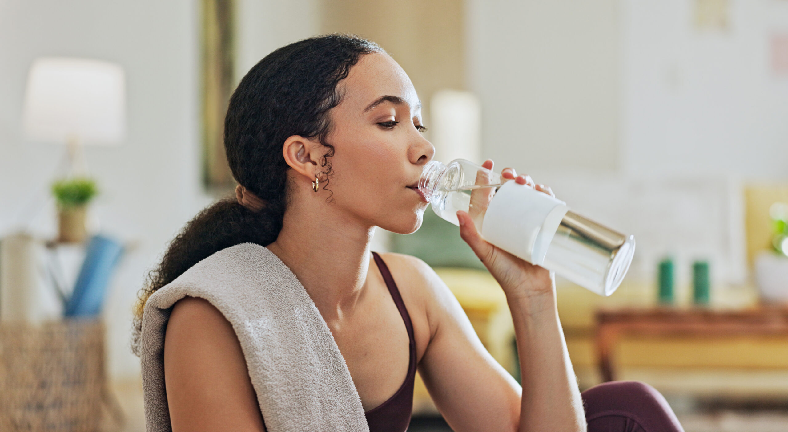A young woman shown from the shoulders-up, with a sweat towel draped over one shoulder, drinks from a large water bottle after a tough workout.