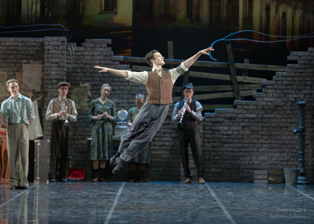 Joel Calstar-Fisher does a cabriole derriere onstage, wearing gray slacks, a cream button-down, and a sepia textured vest. 