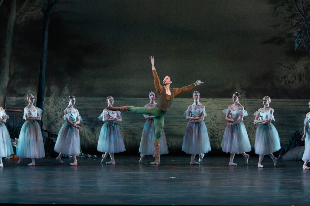 In front of a line of dancers dressed as Wilis, Fernandez jumps. He wears green tights and a brown tunic and vest.