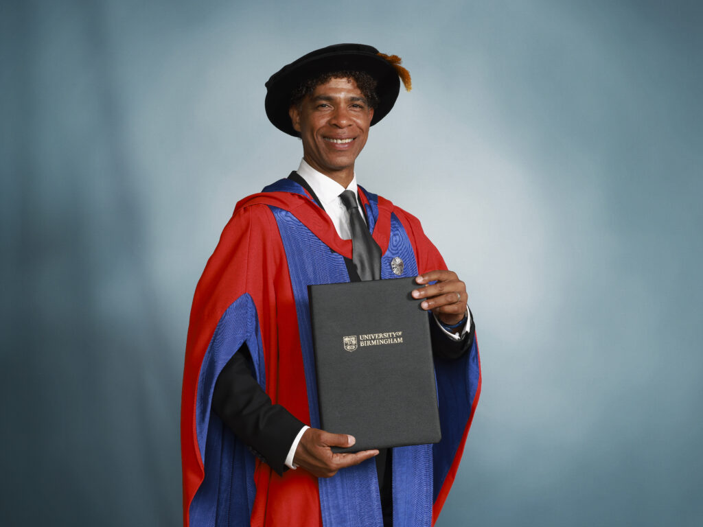 Carlos Acosta, in academic dress, poses in front of a light blue backdrop holding an honorary degree.