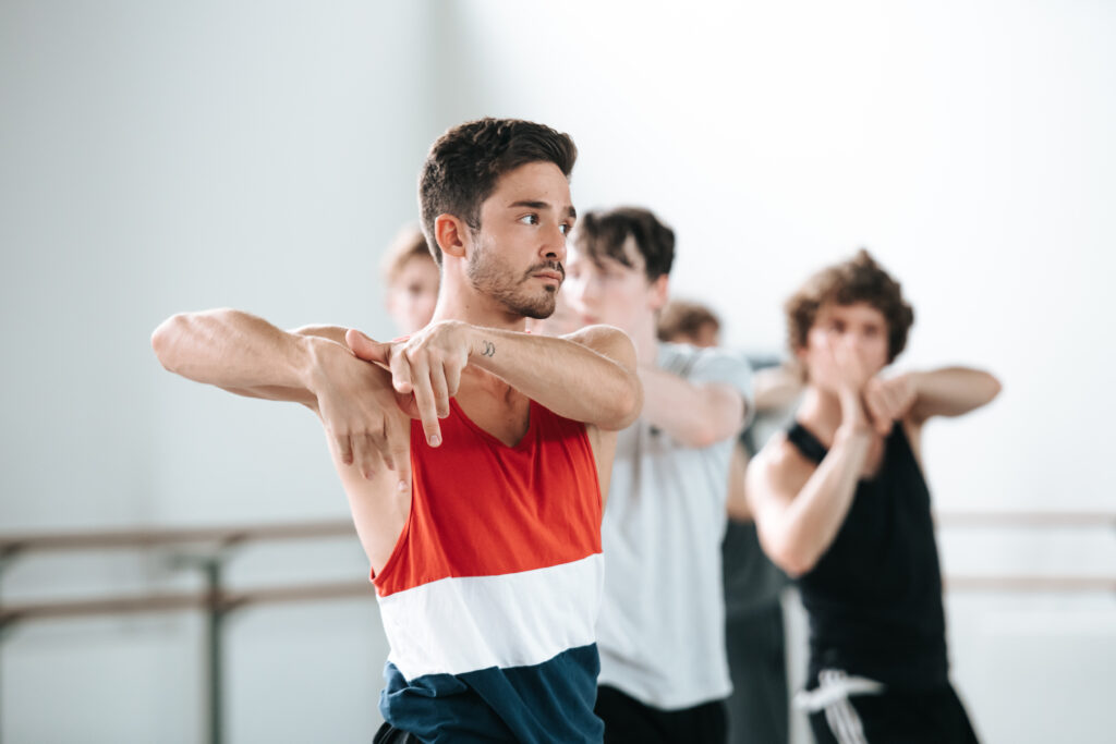 In a brightly lit studio, Javier Andreu (and other dancers behind him) rehearse a contemporary ballet. They bent their elbows up and out to either side, twisting so their fingers point down in front of the right side of their chests.
