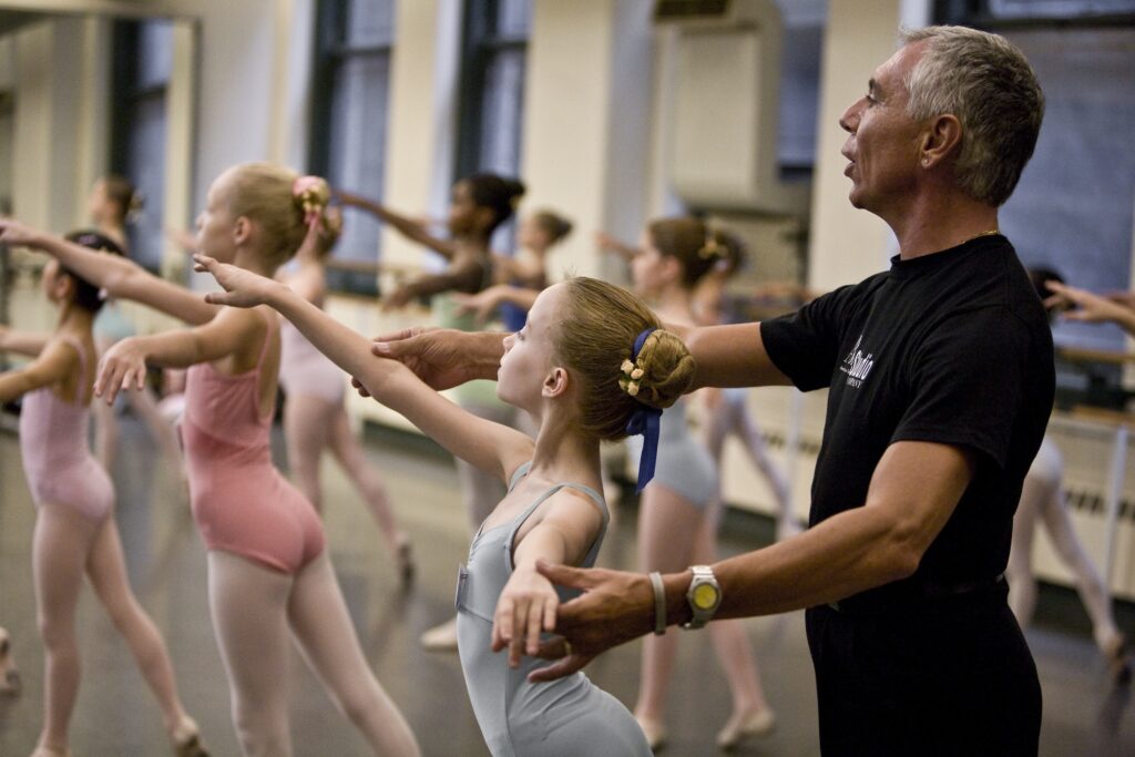 Franco De Vita, wearing a black t-shirt and pants, stands behind a young ballet student as she does tendu croisé derriere with her arms in first arabesque. He holds her forearms to make sure they are in the proper position.