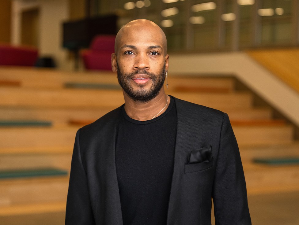 Antonio Douthit-Boyd, shown midsection-up, poses for a headshot in front of a staircase. He wears a black shirt under a suit jacket and a long earring.