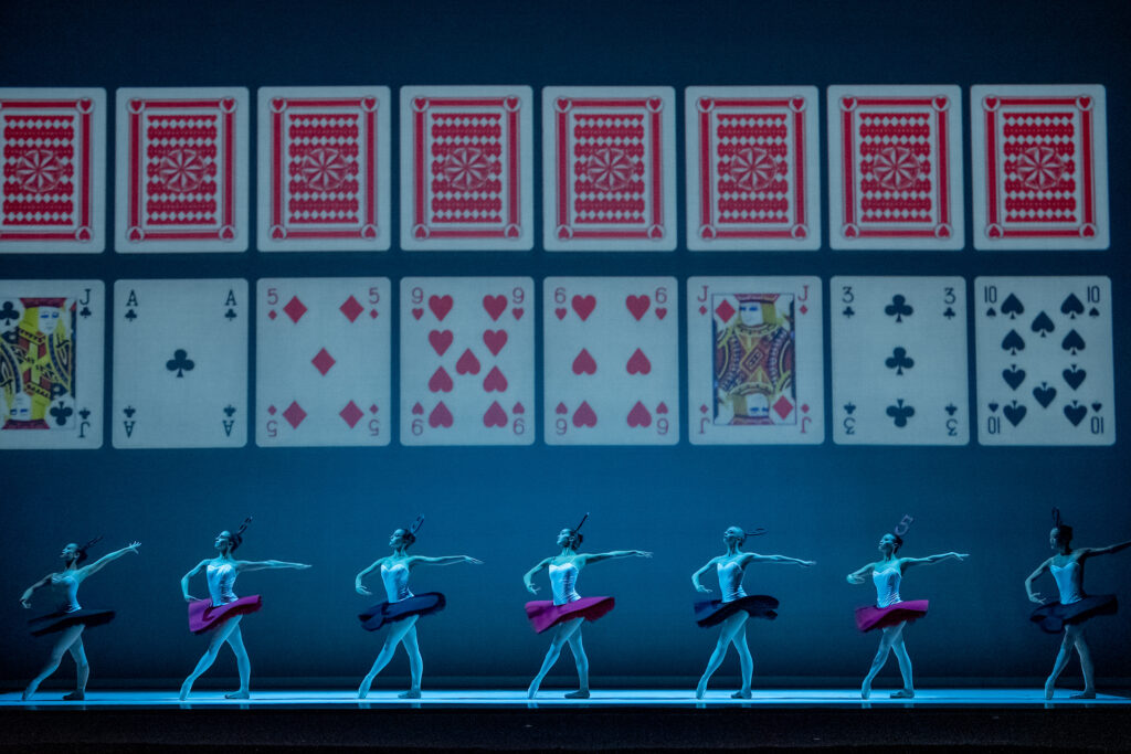 Seven ballerinas pose in tendu croisé devant onstage under cool blue lighting. They wear tutus with white bodices and either red or black tutus, pink tights and pointe shoes. Above them is a large projection showing two rows of playing cards.