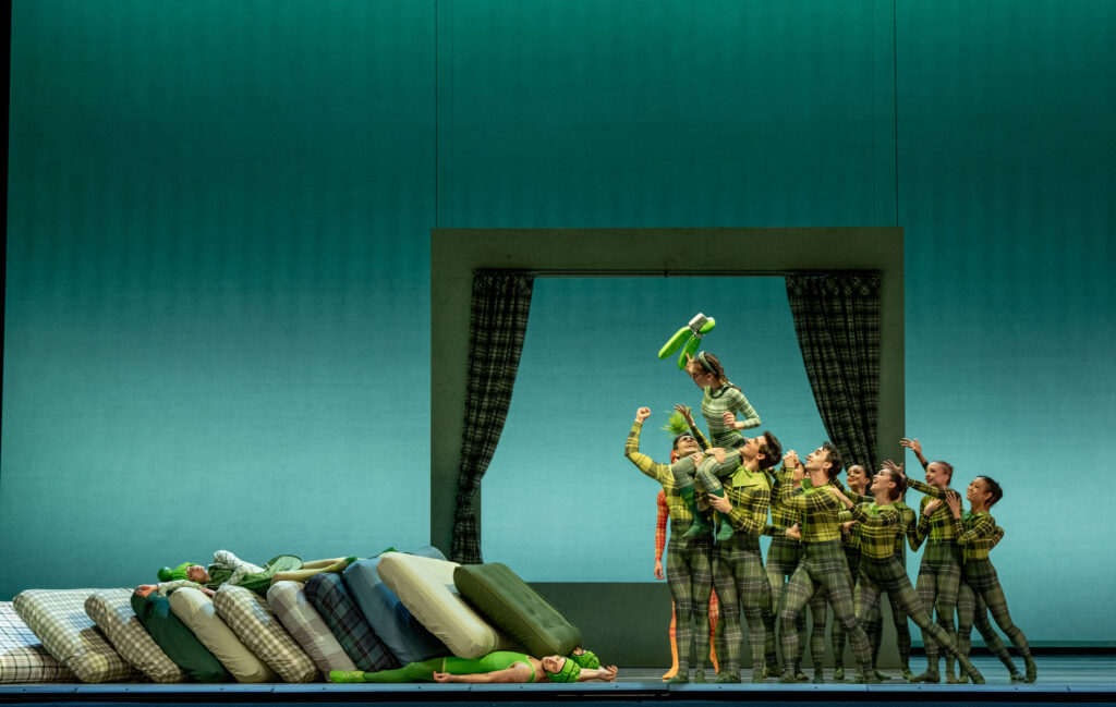 A group of dancers in green plaid unitards holds up another dancer on their shoulders as she lifts a green object above her head. They all look up at her and smile. To their right is a horizontal pile of mattresses, with two dancers sleeping underneath and one lying on top.