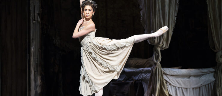 Yasmine Naghdi does an arabesque onstage. She lifts her right arm and crosses her left arm across her chest and resting her left hand on her right shoulder. She is costumed in an off-white dress with lace trimmings, pink tights, and pointe shoes, along with a bejeweled headpiece and bracelet.