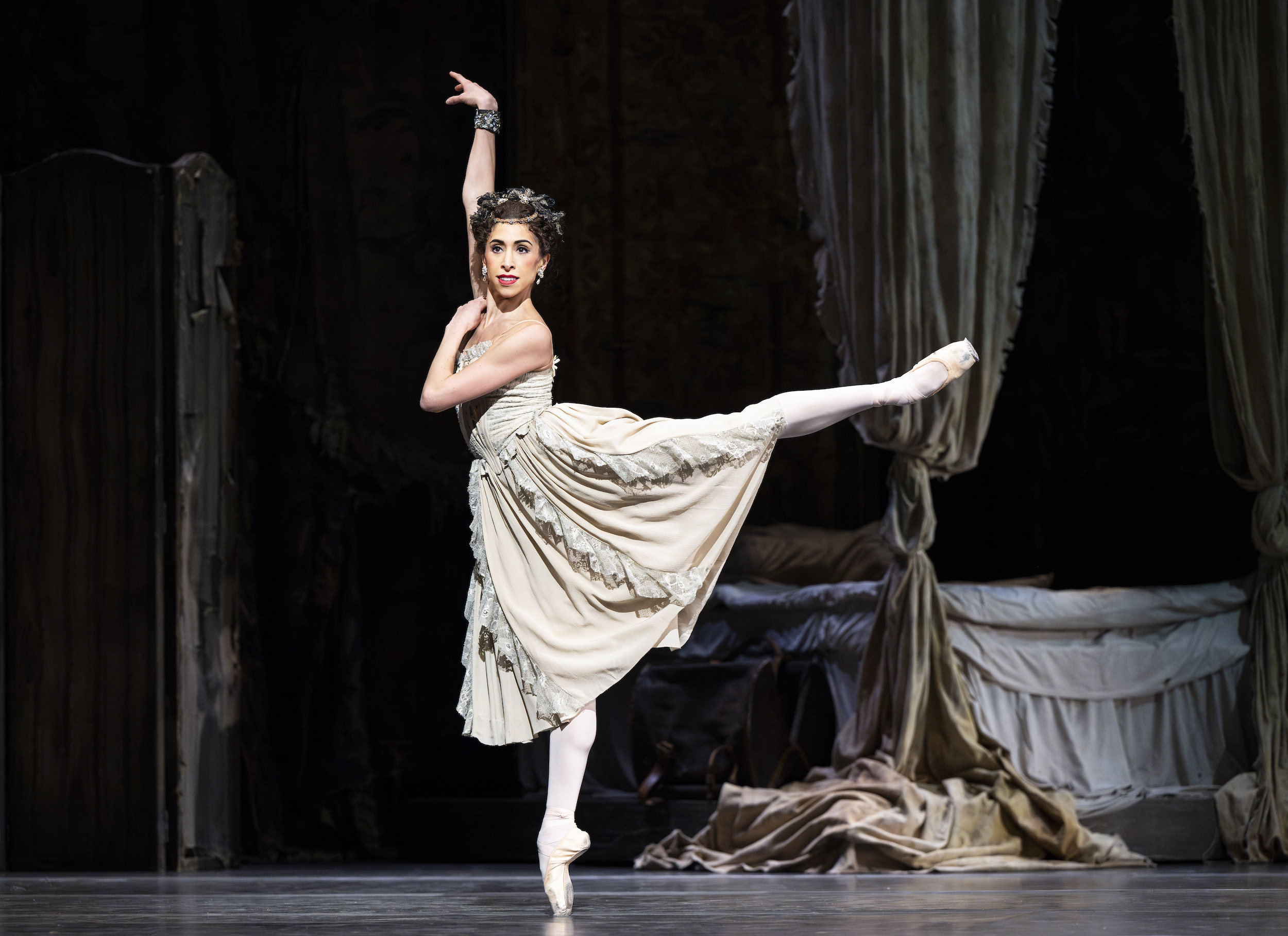 Yasmine Naghdi does an arabesque onstage. She lifts her right arm and crosses her left arm across her chest and resting her left hand on her right shoulder. She is costumed in an off-white dress with lace trimmings, pink tights, and pointe shoes, along with a bejeweled headpiece and bracelet.