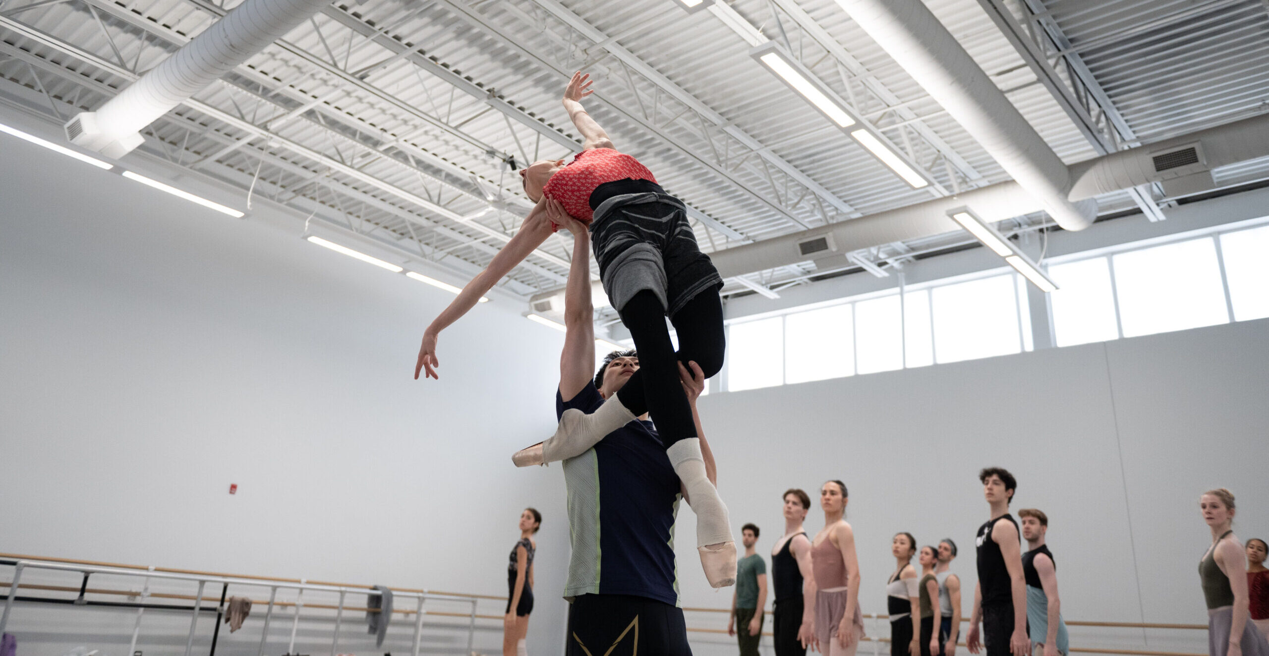 A male dancer holds a female dance in a lift as she extends backwards with her arms in an L shape, holding her left leg in a low coupe attitude back.