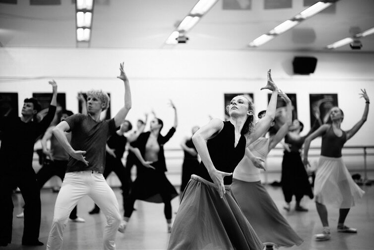 A group of dancers wearing skirts and pointe shoes lean away from the camera with left arms curving up behind their heads while right arms are turned in curving in front of the body.