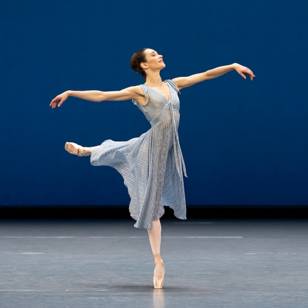 A dancer wearing a light blue dress poses en pointe in attitude against a dark blue backdrop.