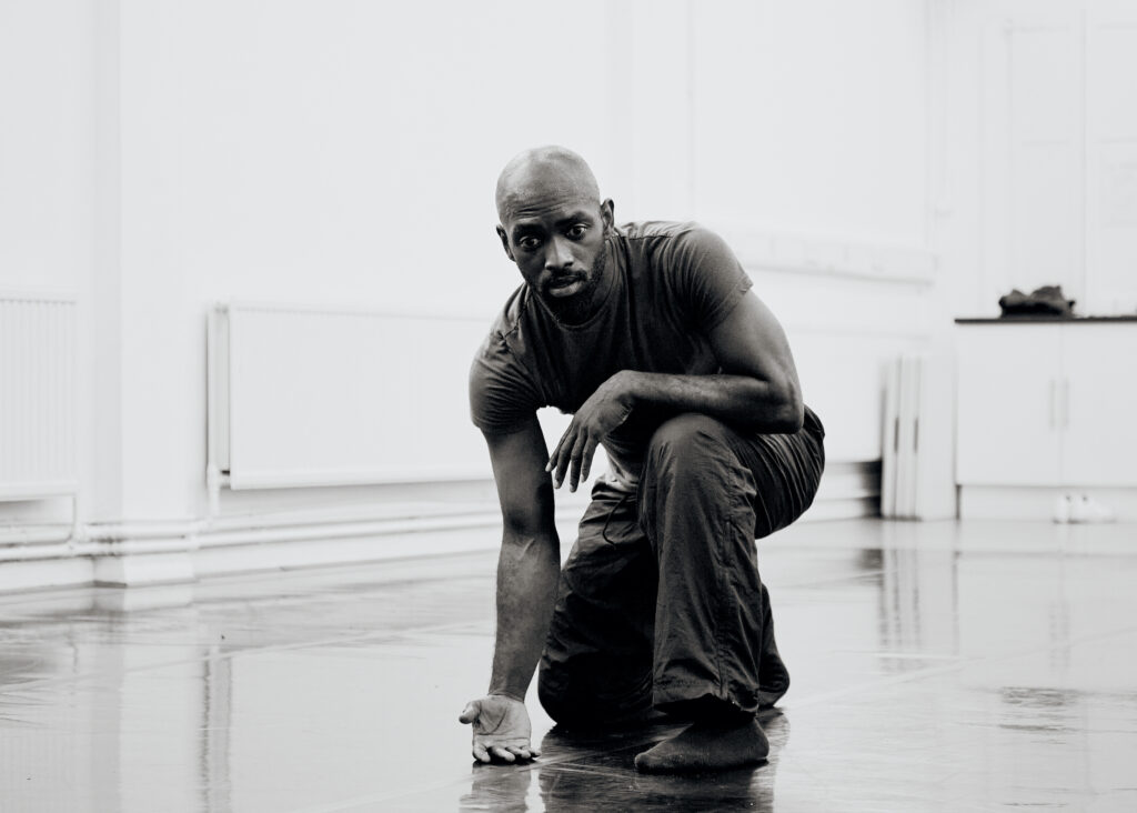 In a brightly lit dance studio, Dickson Mbi kneels down onto his right knee, resting his left arm across his left thigh and touching the floor with the back of his right hand. He wears a dark t-shirt, workout pants, and dark socks,