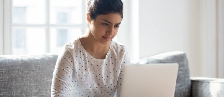A young woman sitting in a chair focuses on the screen of her laptop while typing. The laptop rests on a tabletop.