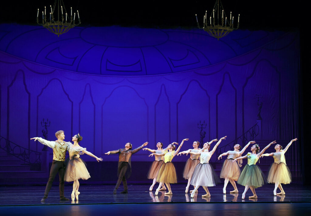 A group if seven women and one man pose in tendu croisé, their standing leg in plié, as they lean back slightly and extend their arms long. The women wear colorful knee-length tutus, pink tights and pointe shoes, while the danseur wears a brown tunic and black pants. On stage right, a centra couple in yellow pose and look at each other, the woman in sous-sus and her partner holding her by the waist.