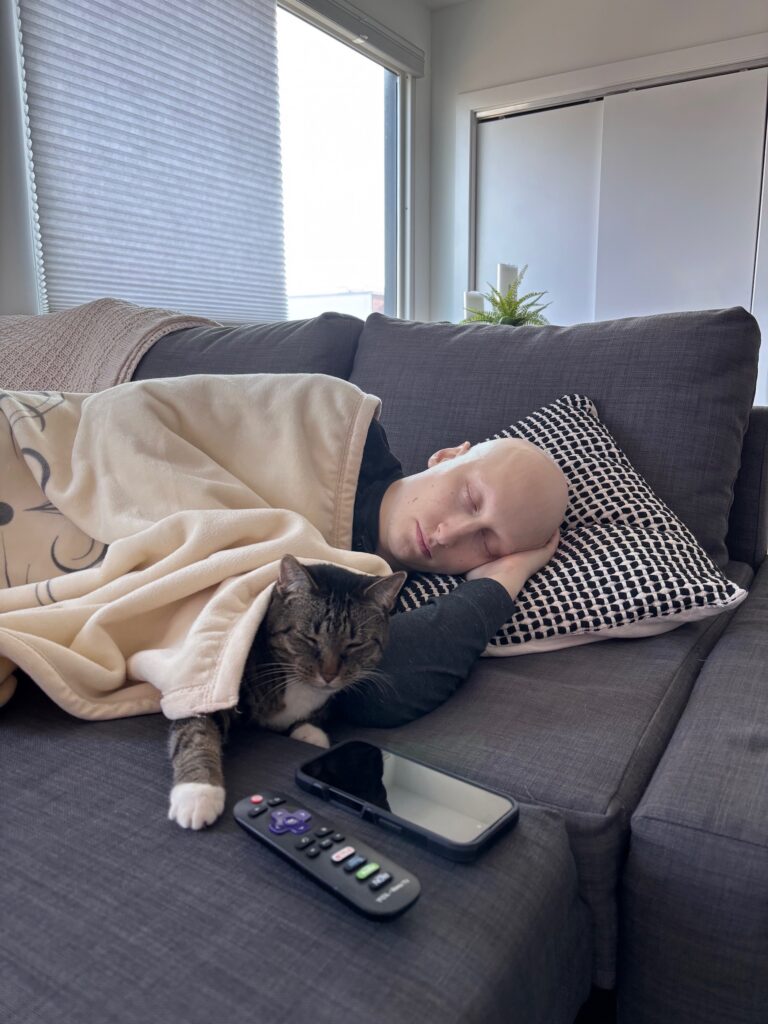 A bald man sleeps on his couch, covered by a yellow blanket, alongside his cat.