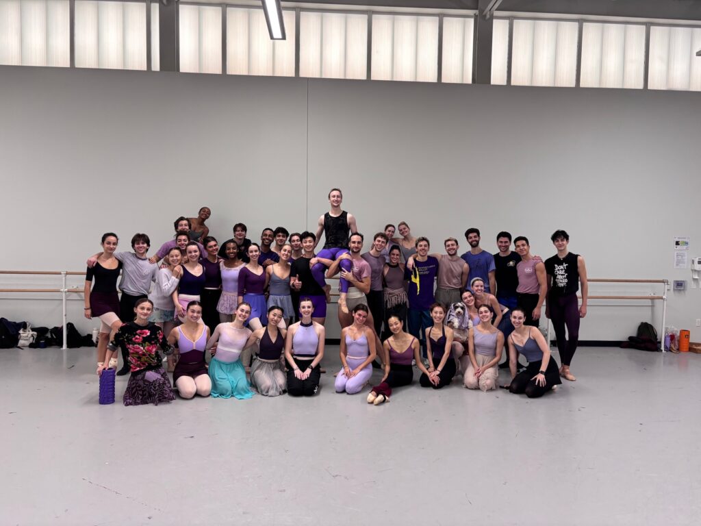 A large group of dancers pose for a photo, with the back line standing and the front line kneeling. They all wear purple practice clothes. A male dancer in the center is lifted onto two dancers' shoulders.