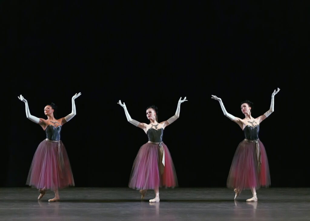 Three ballerinas pose in b plus with their arms lifted, elbows bent and wrists flexed. They wear long pink and black tutus with black bodices and long white gloves. They tilt their heads slightly to their right, looking out at the audience.