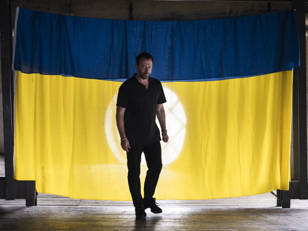 A man wearing black poses in front of a blue and yellow backdrop resembling the Ukrainian flag.