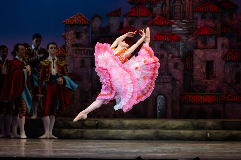 Costumed in a white, Spanish-style dress with pink and orange frills, Meilssa Gelfin does a large sissone with her back leg in attitude. She arching back, almost touching her head to her foot, with her arms in high fifth. She dances in front of a Spanish village set piece, with fellow dancers in costumes watching in the background.