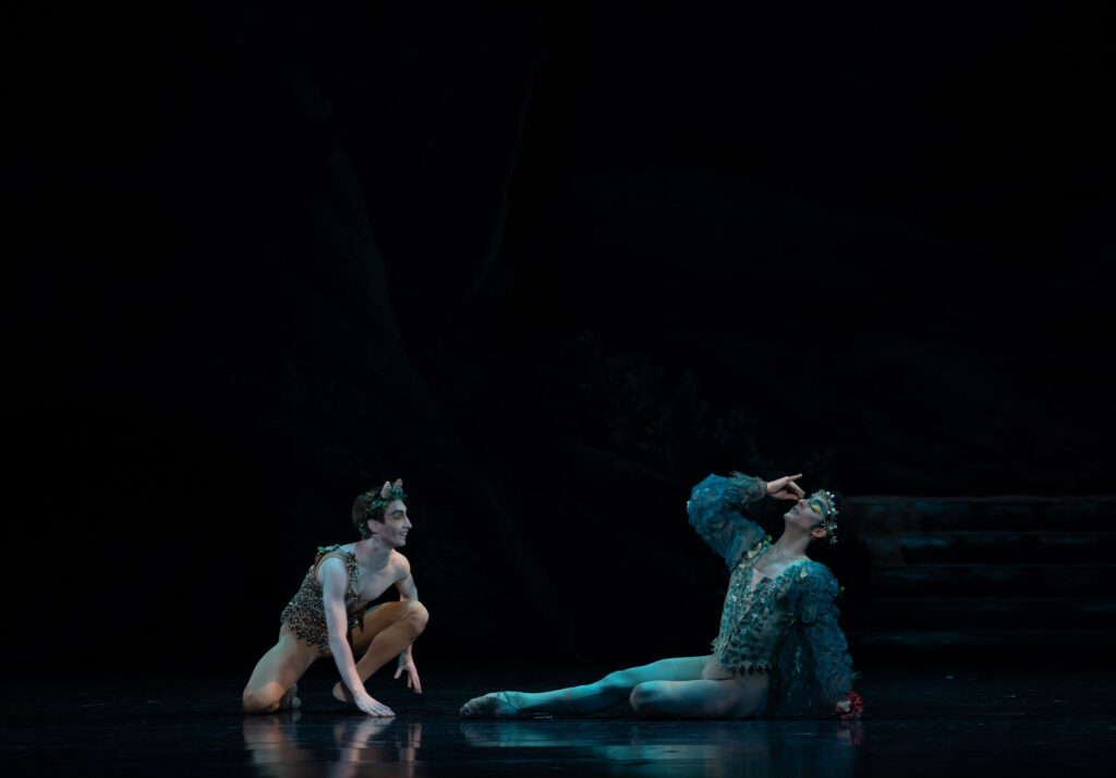 Nicholas Patterson, wearing a brown tunic and tights and a wreath of leaves around his head, kneels down and smiles at another male dancer on his left, who wears a green tunic and tights and leans back, touching his eyes.