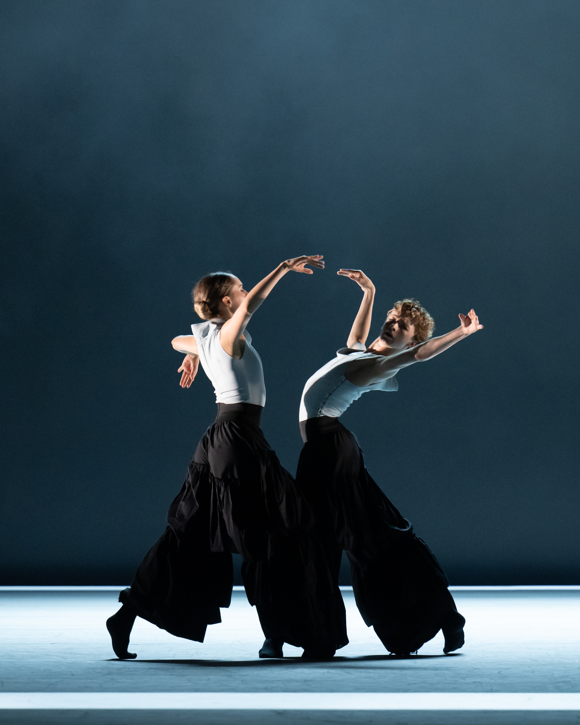 Two dancers in white sleeveless tops and baggy black, wide leg pants stand facing one another, arching their backs and raising their arms up. They dance in front of a blueish gray backdrop.