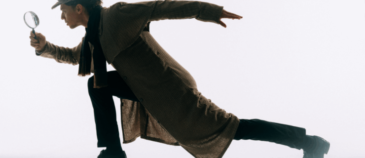 A male dancer wearing a black scarf, plaid cap, and matching plaid trench coat lunges to the left while holding a magnifying glass in his right hand with his left arm extended behind him.