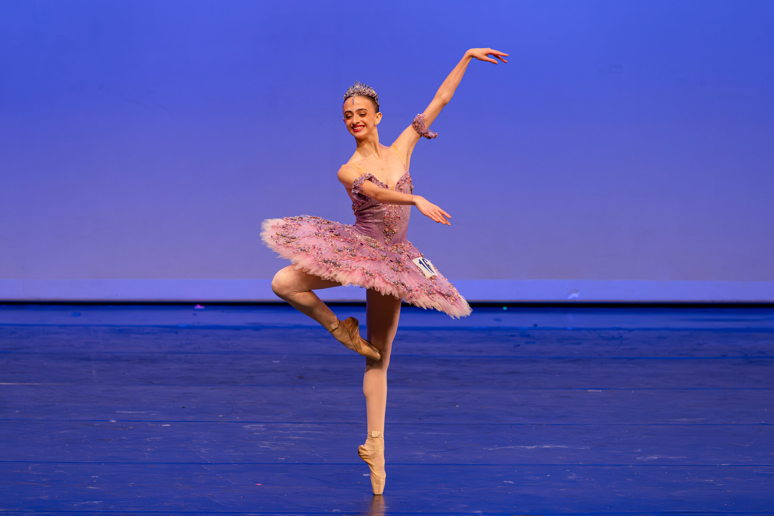 Onstage, Rafaella Pinheiro Marion balances in retiré on pointe with her arms in the third arabesque position. She wears a purple tutu with pink flower bud details and a matching tiara, and a number is pinned onto her tutu.