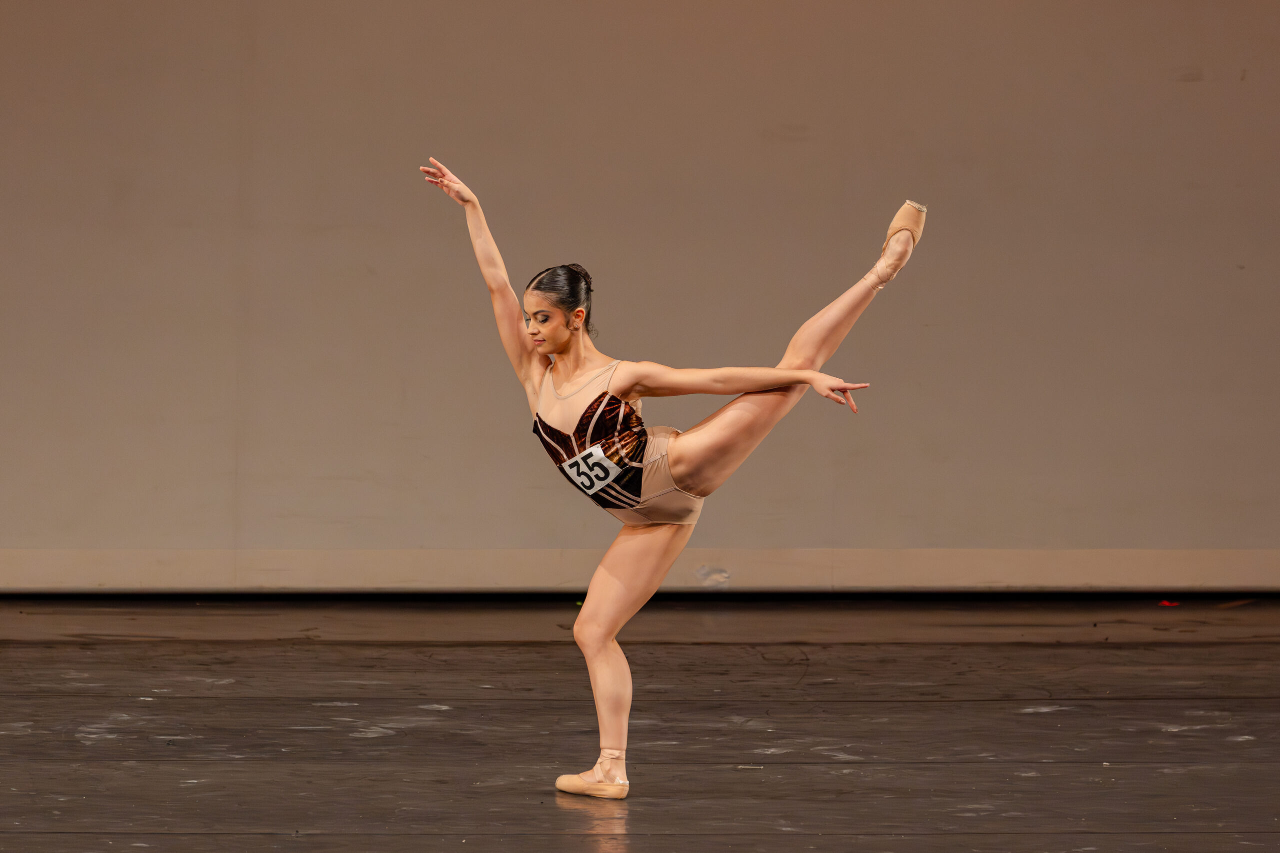 Onstage, Samantha Maestre Cortez poses in arabesque with her bottom leg in plié as she looks slightly down, her arms extended in a high arabesque position. She wears a reddish-brown leotard with nude mesh at the top and a nude-gold bottom.