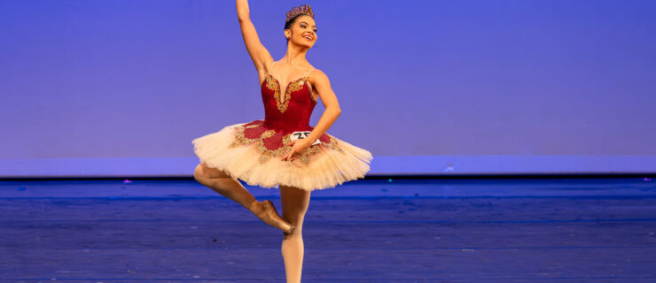 Onstage, Samantha Maestre Cortez balances in retiré on pointe with her arms in high fourth. She wears a red and gold tutu with gold detailing and a matching tiara, and a number is pinned onto her tutu.