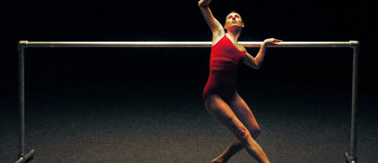 Onstage, Tiler Peck poses in tendu croisé devant with her supporting leg bent, looking up at her top arm, which bends at the elbow and wrist. Her other arm rests on a barre behind her. She wears a bright red camisole leotard.