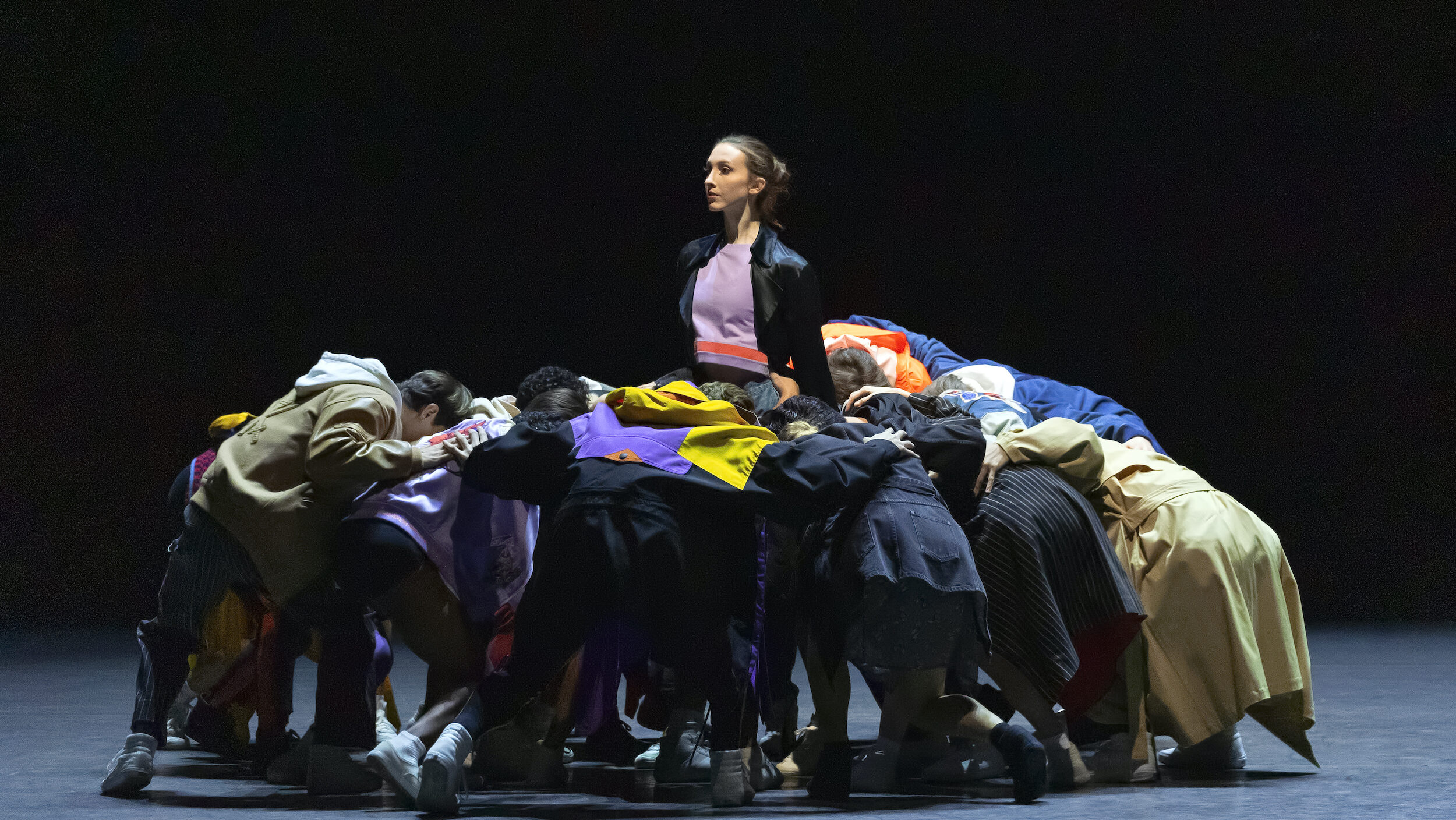 A lone woman in a black jacket and purple shirt stands onstgae, looking towards stage right, as a group of dancers surround her, standing close together and bent at the waist.