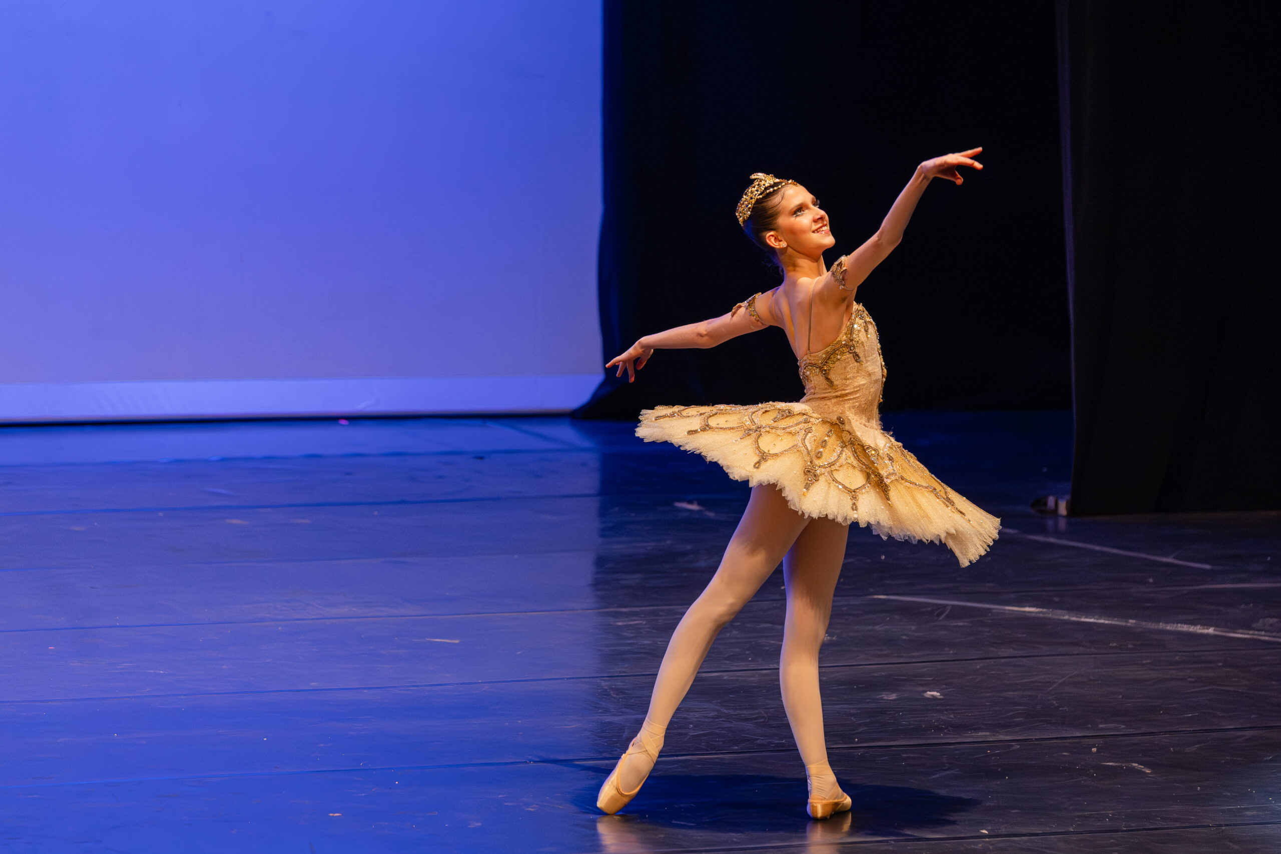 Onstage, Valentina Toscani poses in tendu derriere with her torso slightly twisted so her extended arms match the line of her back leg. She wears a gold tutu with gold detailing and a matching tiara.