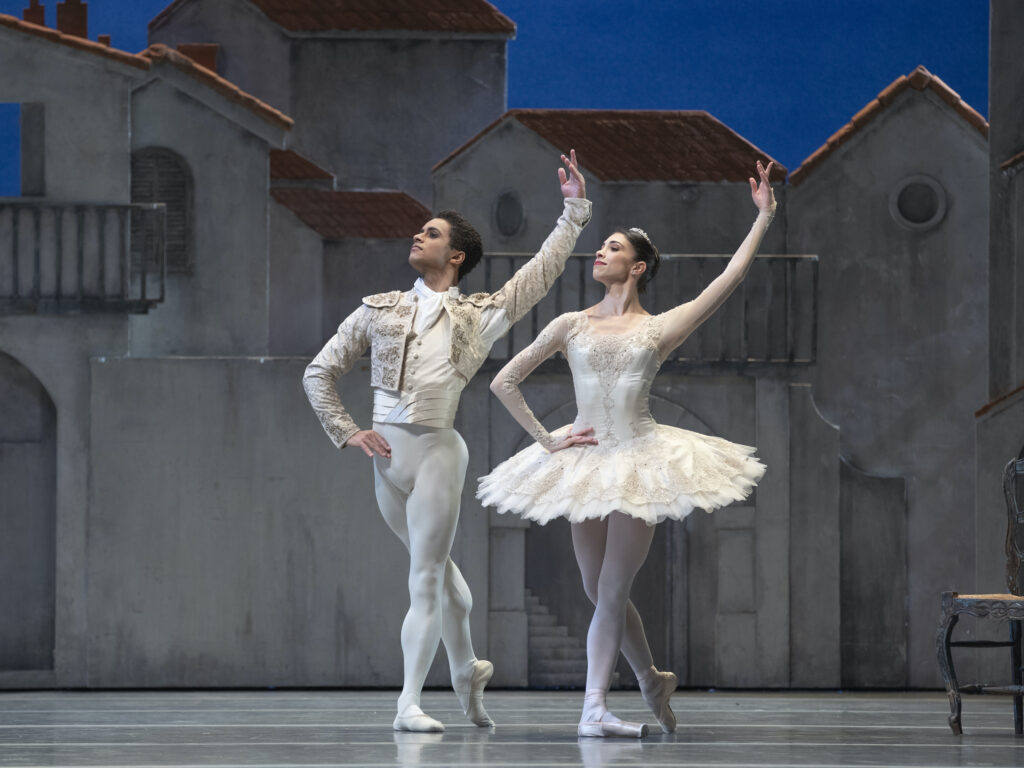 A male and female dancer pose in B plus onstage, with their right hand on their hip and their left arm raised above their head. The each wear white costumes with gold trim, with the man in a bolero jacket and white tights and the woman in a white classical tutu, pink tights, and pointe shoes.